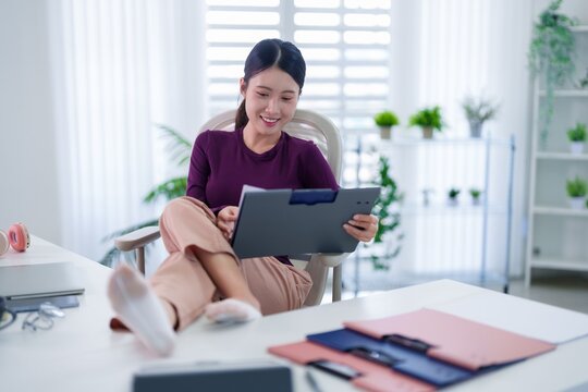 happy Asian women using tablet computer sitting chair relaxing feet on table remote work home office space with plants bright window comfortable casual