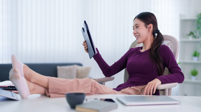 Asian woman freelancer using tablet computer for video call relaxing with feet up at home office desk enjoying comfortable hybrid work lifestyle remote working