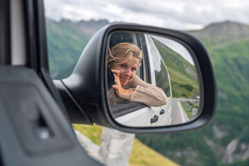 Obraz premium A young woman is seen in the vehicle side mirror as she enjoys a road trip through the Alps, capturing the feeling of freedom, adventure, and the joy of travel against scenic.