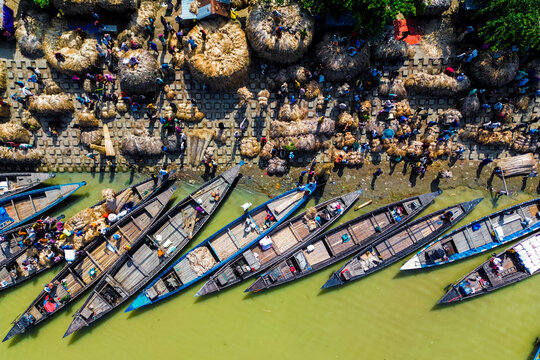 Islampur, Bangladesh - 05 September 2025: Aerial view of traditional wooden boats docked at a busy riverbank market with workers moving large bundles of jute under bright sunlight.