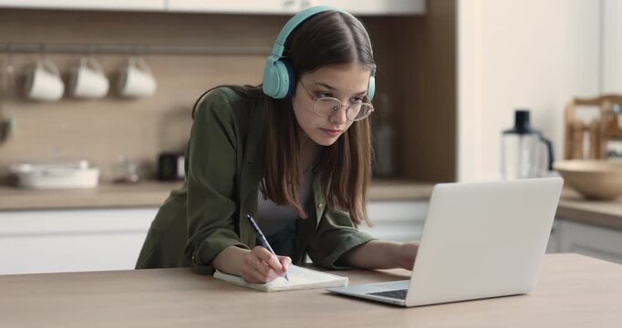 Teen girl wears headphones listening audio course, studying, making exercise, writing notes in copybook, preparing homework, e-learning using laptop in domestic kitchen. Online education from home