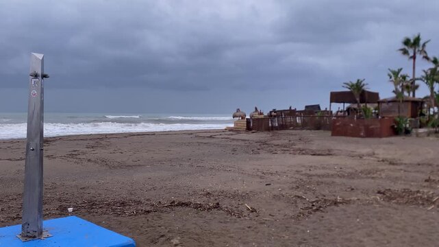 Empty beach shower standing on a dark sand beach with rough ocean waves and an abandoned beach bar under cloudy skies