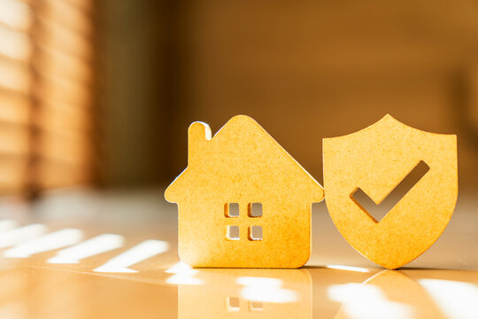 Wooden house model standing next to shield icon with check mark on sunlit floor, concept of home insurance, property protection, legal safety, residential security and asset guarantee.