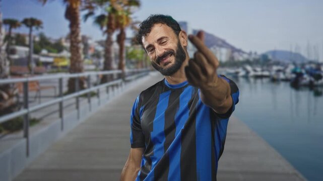 Man in soccer jersey shows middle finger while smiling on a street side marina pier with palm trees and boats; defiance.