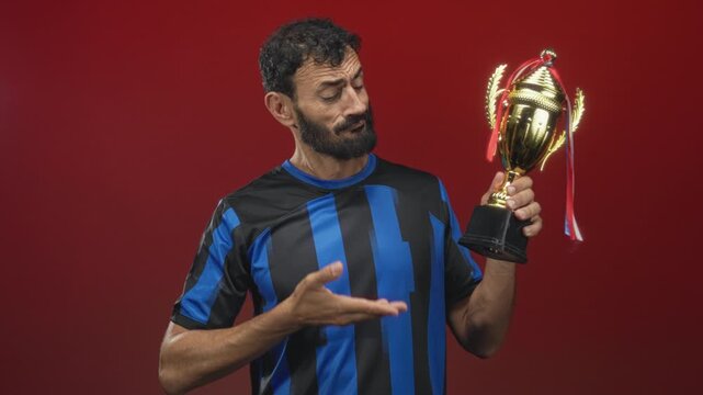 Man in soccer uniform points finger at trophy while holding cup against red studio backdrop; victory pride.
