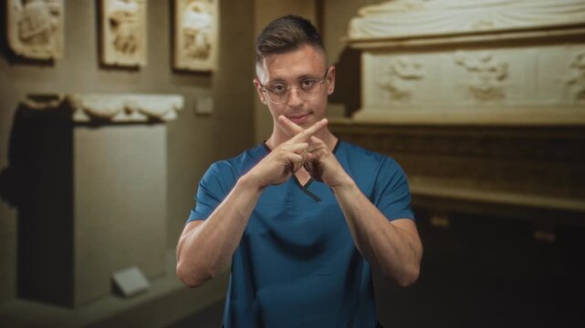 Doctor man in blue scrubs and glasses crossing forefingers into an x with hands, standing in a museum building gallery near stone sarcophagus and sculptures; denial caution.