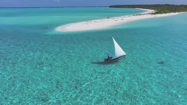 Aerial view of a traditional sailboat with a white sail gliding through turquoise waters near a white sandbank in Bileffahi, Shaviyani Atoll, Maldives.