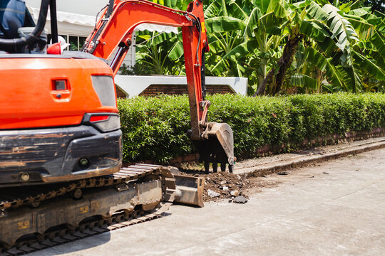 An orange crawler excavator digs a trench along a paved residential road for maintenance.