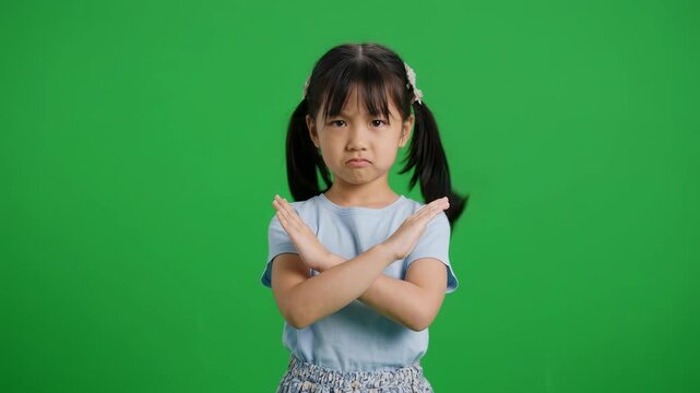 Cute Asian little girl making a stop or no gesture by crossing arms in X sign on green screen background for refusal disagreement and safety warning concept