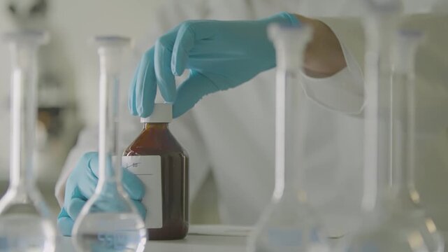 Scientist with blue gloves handles brown reagent bottle among glass lab flasks