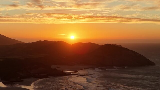 A sweeping aerial view of the New Zealand South Island coastline at peak sunset, capturing the intense golden glow over the mountains and the rhythmic waves washing onto the shore.
