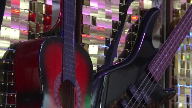 A guitar and electric bass sit side by side in a music shop. The sun shines through the window, casting light on the instruments. Customers explore the shop and check out various music gear.