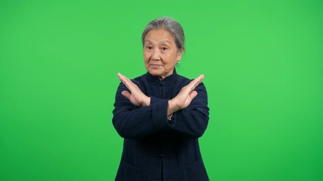 Senior Asian woman making an X gesture with her arms to express refusal or prohibition for health warnings and safety social media content on green screen background