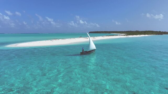 Aerial view of a traditional dhoni sailboat with a white sail gliding through turquoise waters near a tropical island with white sand in Bileffahi, Shaviyani Atoll, Maldives.