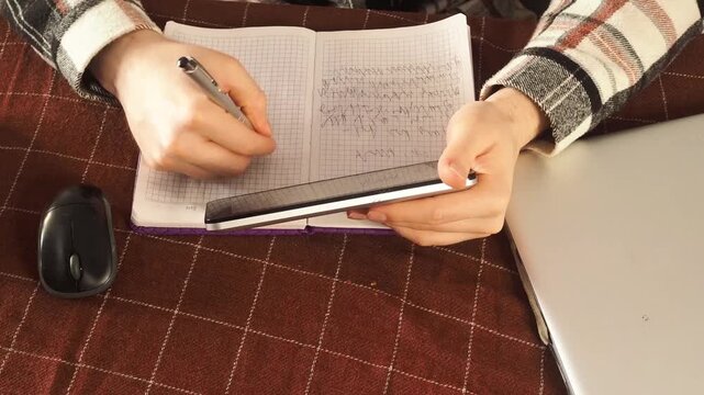 Close-up hands of male student writing notes with pen on notebook, reading book sitting at table in dark home setting.