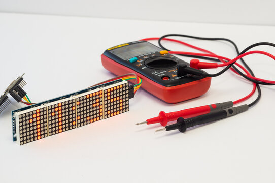 A multimeter with test probes and a Wi-Fi clock with seven-segment displays and ESP32 lie on a white background.
