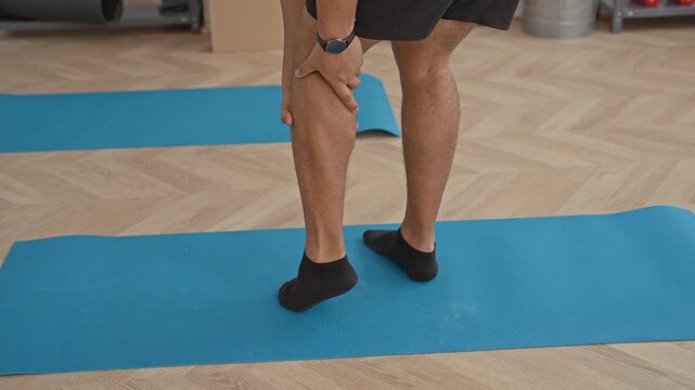 Young man grips calf muscle with both hands while standing on a blue exercise mat in studio during a training session; pain recovery rehab.