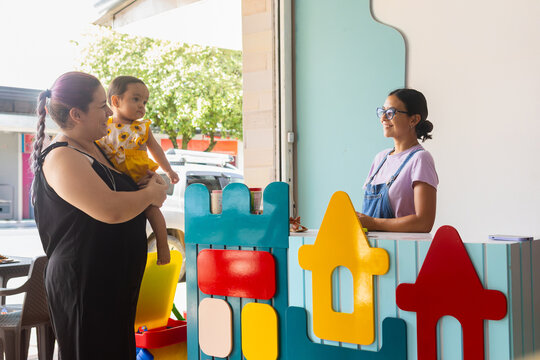 Two adult women and a baby entering a colorful early childhood stimulation center, reflecting care, early education and family support in a learning environment, baby 1 to 2 years old and adult Latina