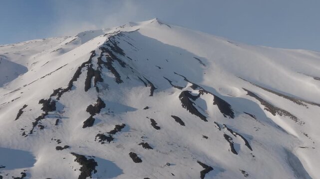 Aerial view of the snow-covered peak of Mount Etna with dark volcanic rock patches under a clear sky near Rifugio Sapienza in Nicolosi, Sicilia, Italy.