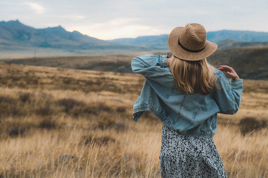 Girl in denim jacket and hat in golden field. Back view with mountains