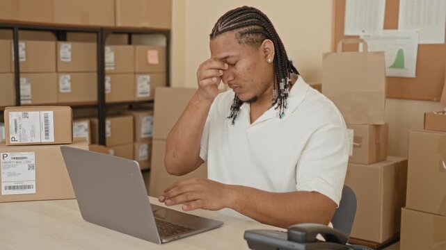 Man puts on glasses and types on laptop among packages in a building packing room; concentration productivity organization.