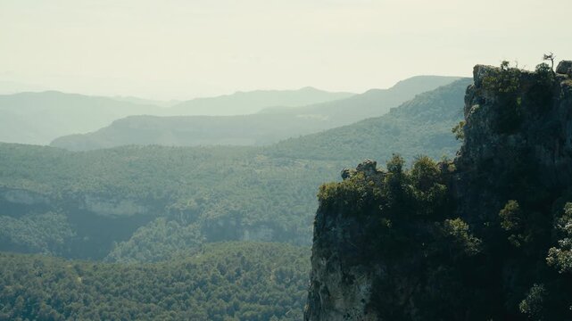 A rugged rock cliff rises above a vast forested landscape, where soft haze and layered ridges create a calm mountain view with depth and dramatic natural scale.