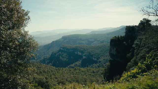 A sweeping view opens across a deep forested canyon with steep cliffs, layered ridges, and hazy distance, creating a dramatic natural landscape with strong depth and scale.