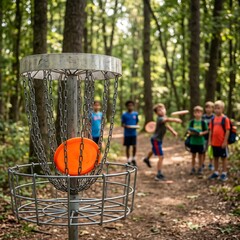 Fototapeta premium A vibrant orange disc landing in a metal disc golf basket in a wooded park, with children throwing in the blurred background, highlighting youthful recreation 1:1 ratio...