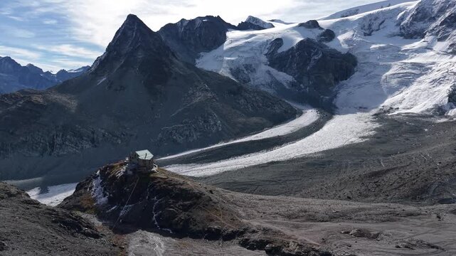 Aerial flyover around mountain hut alongside Glacier de Cheilon as the morning sun shines over the tall mountain peaks of Swiss Alps in the Arolla region