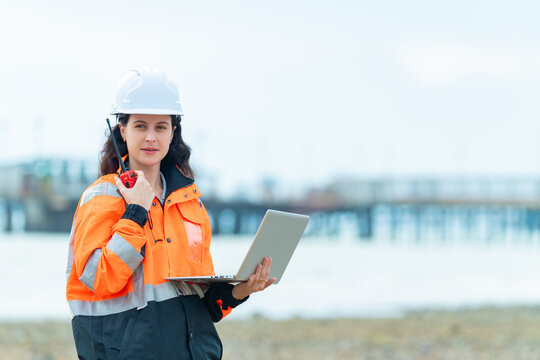 Female industrial engineer using walkie talkie and laptop computer at port facility, professional logistics worker in safety helmet, transportation industry communication and cargo terminal operations