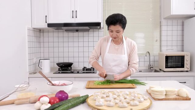 Portrait of elderly woman baker chef chopping fresh chives or cutting green onion on kitchen cutting board with knife to making tasty homemade dumplings stuffed with minced meat and vegetable for buns