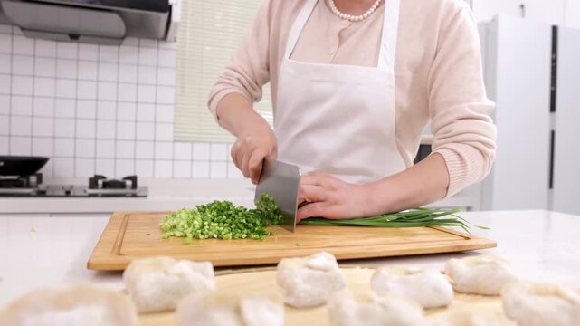 Elderly woman baker chef chopping fresh chives or cutting green onions on a kitchen cutting board with knife to making tasty homemade dumplings stuffed with minced meat and vegetable for filling buns