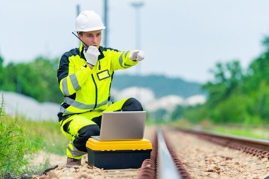 Railway engineer inspecting railroad track with laptop and radio communication device during railway maintenance operation, industrial transportation infrastructure worker checking rail system