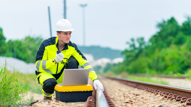 Railway engineer inspecting railroad track with laptop and radio communication device during railway maintenance operation, industrial transportation infrastructure worker checking rail system