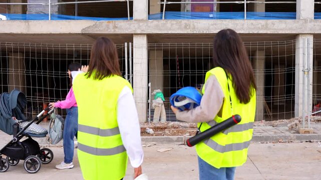 Two female civil engineers discussing building plans at a construction site while a pedestrian passes by with a baby stroller