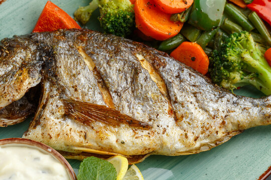 Elegantly Plated Grilled Sea Bream Complemented By Fresh Seasonal Vegetables And Creamy Herb Sauce In Seaside Setting