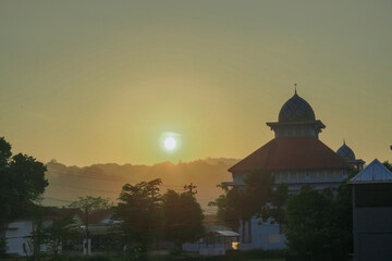 Obraz premium The serene view of the sunrise shows the silhouette of a mosque with a colorful dome standing majestically among the trees and residential areas.