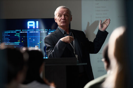 Senior Caucasian businessman presenting AI data during conference talk, gesturing toward screen and audience. Ideal for technology training, corporate communication, innovation marketing