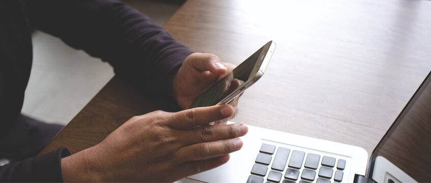 Close-up of hands using smartphone beside laptop on wooden desk, representing online communication, remote work, mobile technology, and digital lifestyle