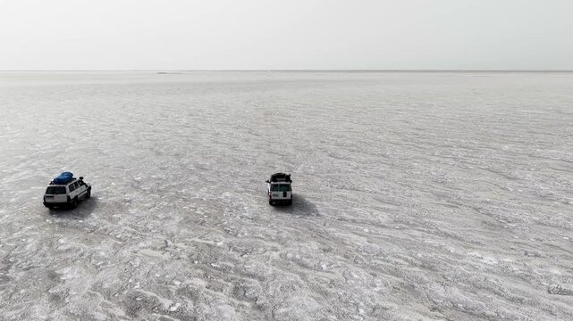 Aerial view of two off-road vehicles driving across the vast white salt crust of the Danakil Desert under a hazy sky in Dalol, Afar, Ethiopia.