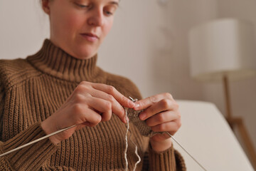 A woman sits quietly at home and uses her hands to knit with a pair of needles. She is focused on her work in a simple indoor space with soft light © sementsova321