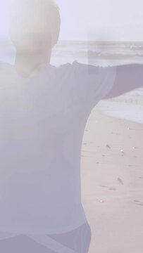 Vertical video: Senior man raising arms running on sand to sea leaving prints sun glare health