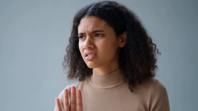 Young woman with curly hair shows stop gesture. She wears beige turtleneck. Her expression changes from calm to frustrated. She clenches teeth and furrows brows. Footage captures refusal, disagreement