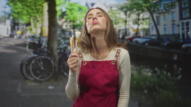Woman in red apron holds three makeup brushes to her face, tilting her head back with closed eyes on a tree lined street beside a canal; serenity.
