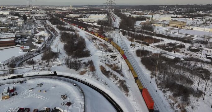Aerial view of a long freight train with colorful containers traveling through a snow-covered industrial area with power lines and bare trees in Reading, Ohio, United States.