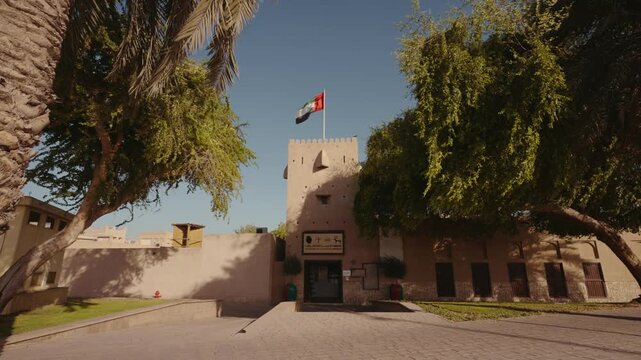 Static shot of traditional adobe fort entrance with flag, framed by palm and green trees under clear blue sky. &middot; 4K DCI 25fps