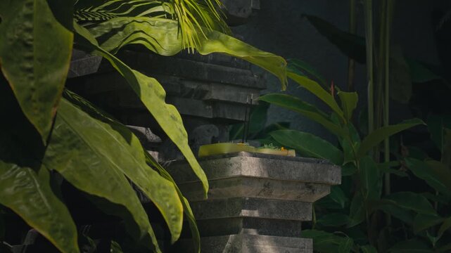Close-up of a traditional Canang Sari offering with smoking incense in a stone temple, surrounded by lush greenery. Peaceful Balinese spiritual ritual with authentic cultural atmosphere.