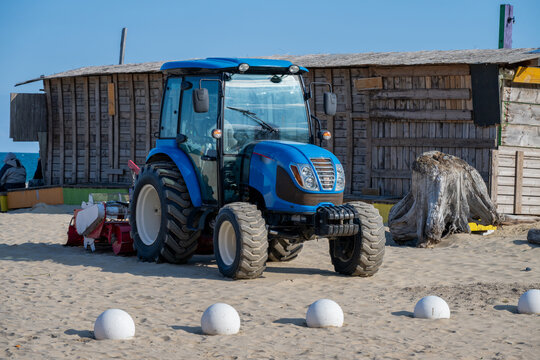 A modern blue tractor with a glass cabin is parked on a sandy beach in front of rustic wooden huts, likely used for shoreline maintenance or beach cleaning.