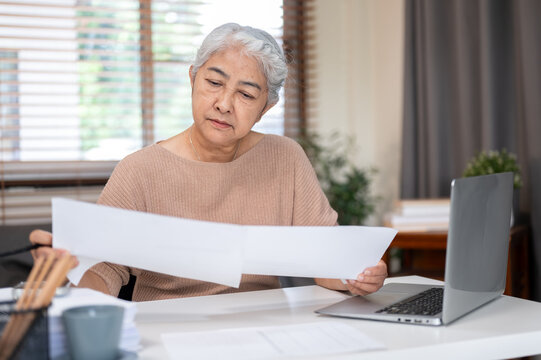 Senior old asian woman holding pencil looking reading paper document on working table at home office