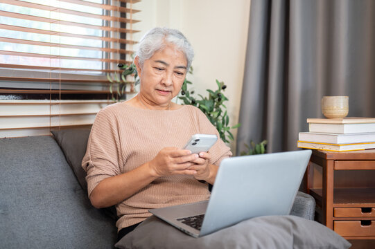 Old asian woman grandma using looking at phone over a laptop sitting on sofa in living room at home.